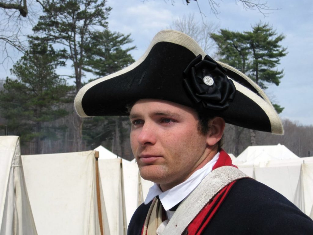 Modern color photograph of a young white man wearing a black cocked had with white trim and a rosette ribbon in black with a silver button at the center. His clothes are a white high-collared shirt, red and blue wool jacket of military design and he has a few white straps in leather and other materials going over both shoulders. In the background is a row of white military tents and further back is a tree line.