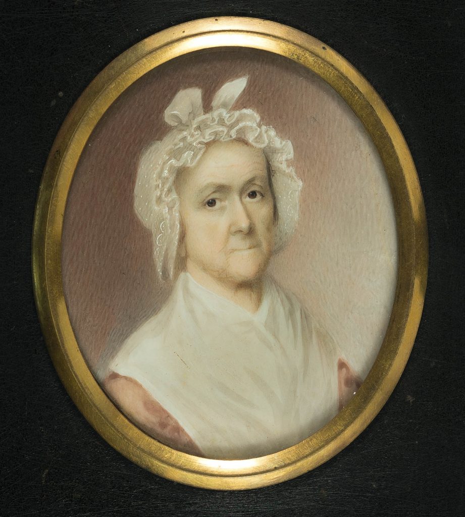 Color photograph of a small portrait painting of an older white woman in an oval gold frame, with black painted wood frame around that. The figure is in ¾ profile and looks at the viewer with expressive dark eyes, she wears a frilled white cap that covers her hair, a large white shawl and a pink dress or shirt. The background is pink, lighter to the right, and gets darker as it goes left.