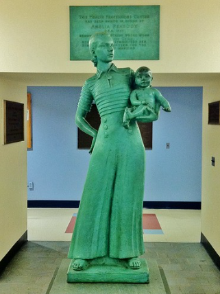 A color photograph of a green metal statue. The statue is a figure of a woman holding a baby on her hip with one arm. She wears long billowy pants with six buttons at the top front and a striped, collared shirt, the stripes are textured. She is in a school building with tiled floors and plaques all around her, including above and behind her head which the viewer can see names the artist, Amelia Peabody, but it is too blurry to read the other words.