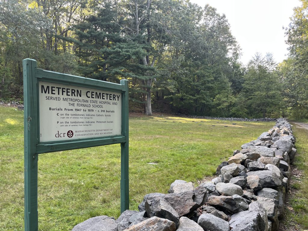 Recent color photograph. In the foreground there is a sign that reads “Metfern Cemetery/Served Metropolitan State Hospital and Fernald School. In the background is a field with some small granite stone grave markers in the ground.
