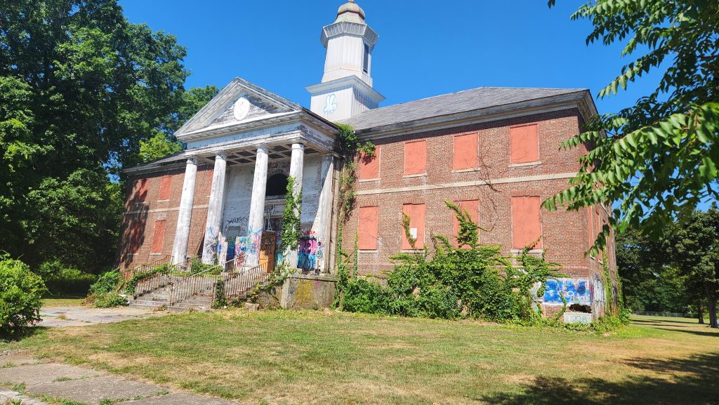 large brick building with white columns in front and a white steeple. The building is surrounded by trees and has graffiti spray painted onto it