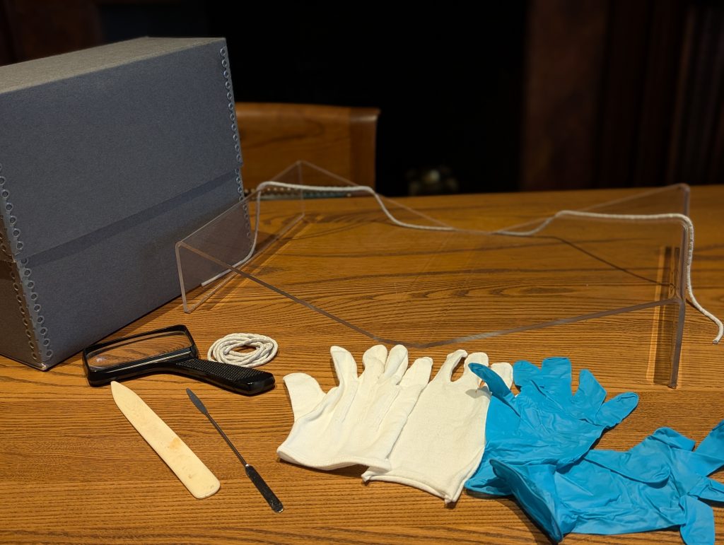 Objects on a table including a manuscript box, book cradle, cloth gloves, latex gloves, and a magnifying glass.