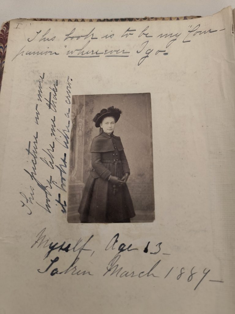 Color photograph of one page of a manuscript volume with some creases and stains. In the center is a black and white photograph of a white girl wearing a dark coat, dark hat, and dark gloves. She is standing, and her fingers are interlocked in front of her. Below the photograph is written, in black and white ink, “Myself, age 13, taken March 1889.” Along the side of the photograph is written, “This picture no more looks like me than it looks like a crow.” At the top of the page is written, “This book is to be my companion wherever I go.”