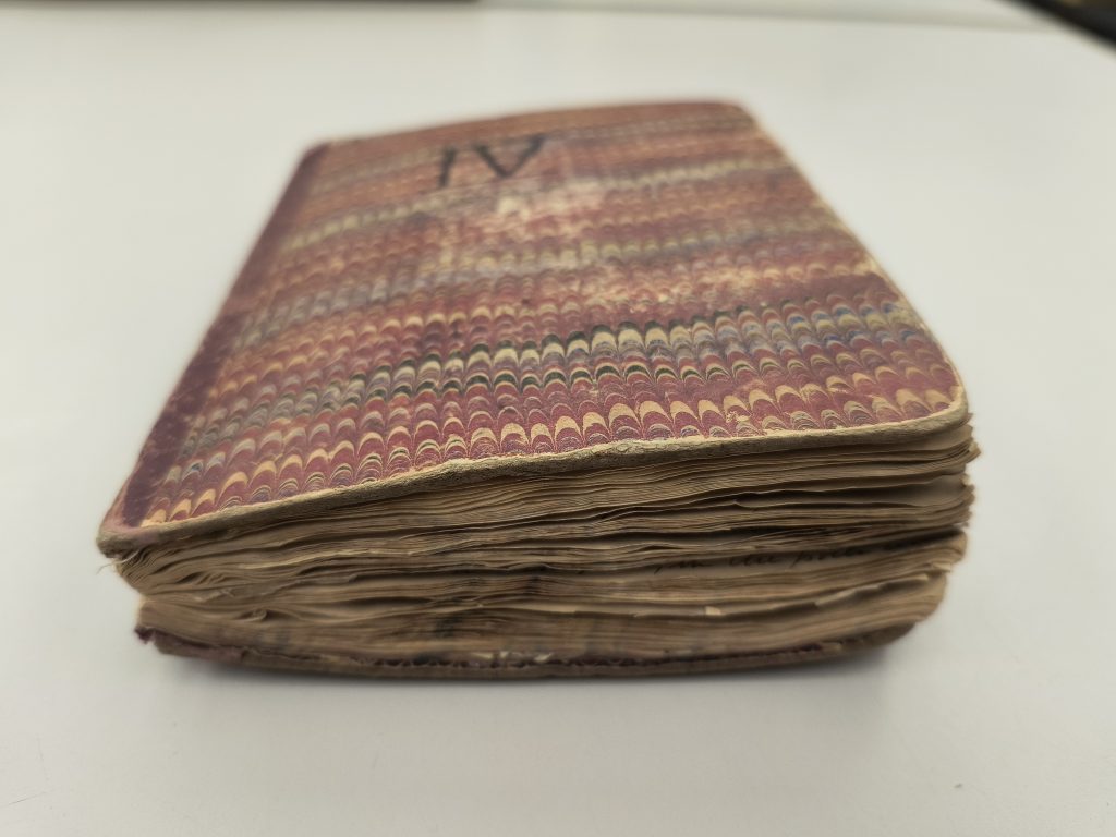 Color photograph of a volume on a table. The cover has a marbled pattern and the letters “IV” written on it in black ink. The pages of the volume are bulging out of its binding.