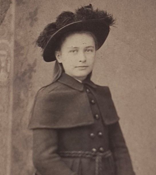 Black and white photograph of a white girl wearing a dark coat, a dark hat decorated with feathers, and straight hair hanging loose down her back.