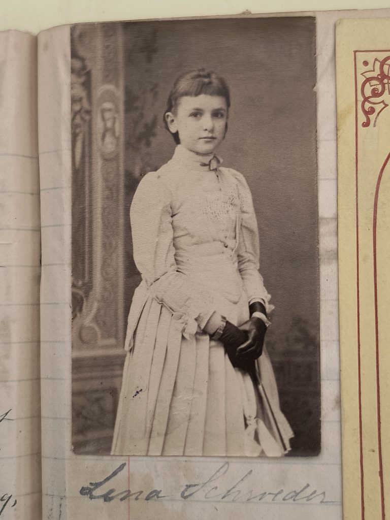 Black and white portrait photograph of a white girl with brown hair in front of a blurry backdrop. She is wearing a white dress and black gloves, and her hands are clasped in front of her. The photograph is pasted onto a lined manuscript page, and below it is written “Lena Schroeder.”