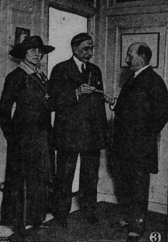 Alt-text: Black and white photograph of a white woman and two white men standing inside a room by a doorway. The white woman wears a wide-brimmed hat, a dark dress, and a medal around her neck. Her hands are clasped behind her back, and she looks directly at the camera. The two men, standing to her left, are smiling at each other and passing an unidentified object between them.
